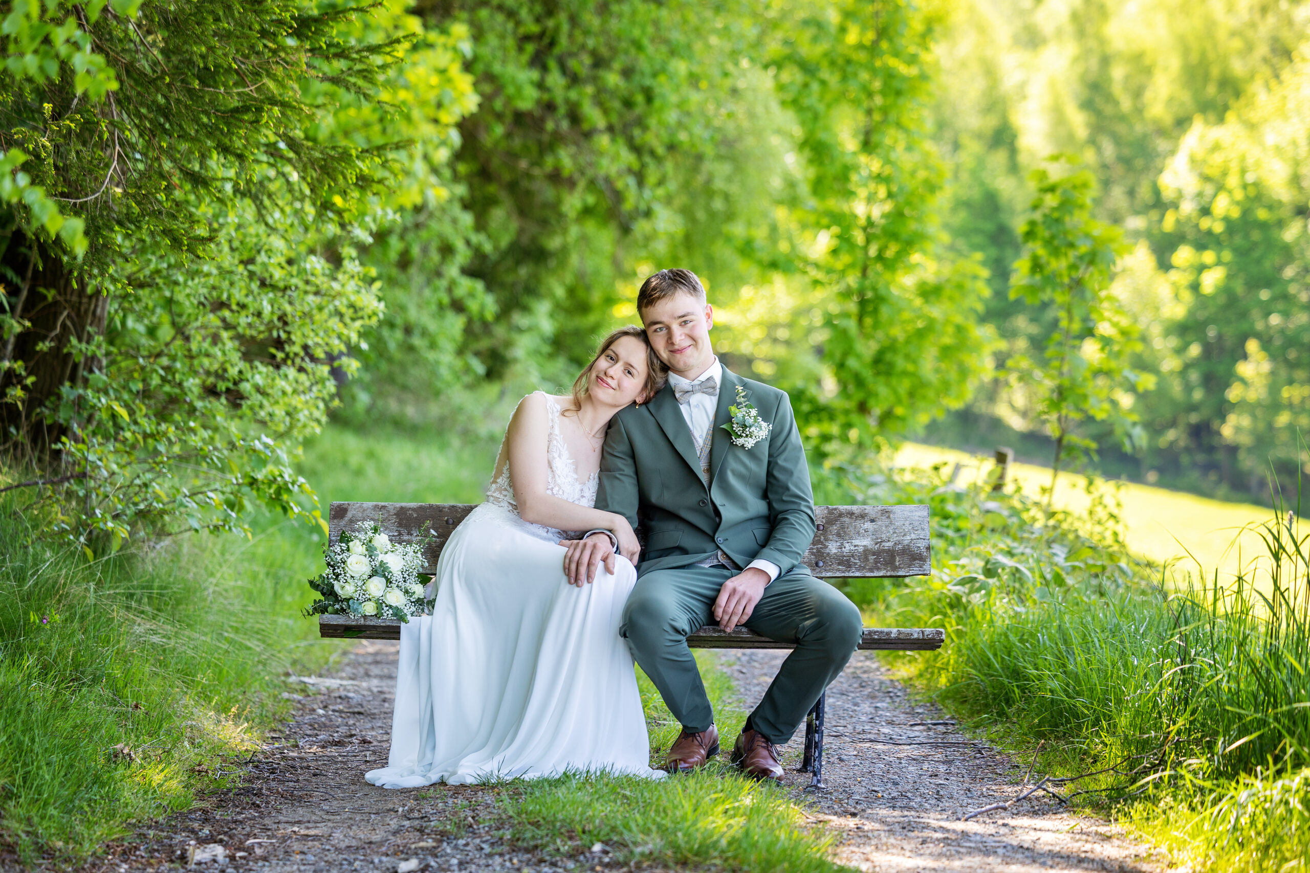 Brautpaar sitzt auf einer Bank am sonnigen Waldweg – Hochzeitsfotograf Marko Lorenz